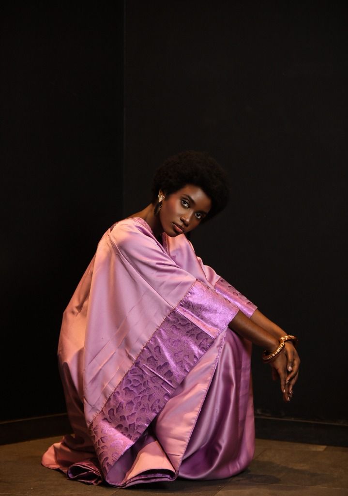 Dorcas Moïra en boubou satin lilas accroupie dans un angle sombre